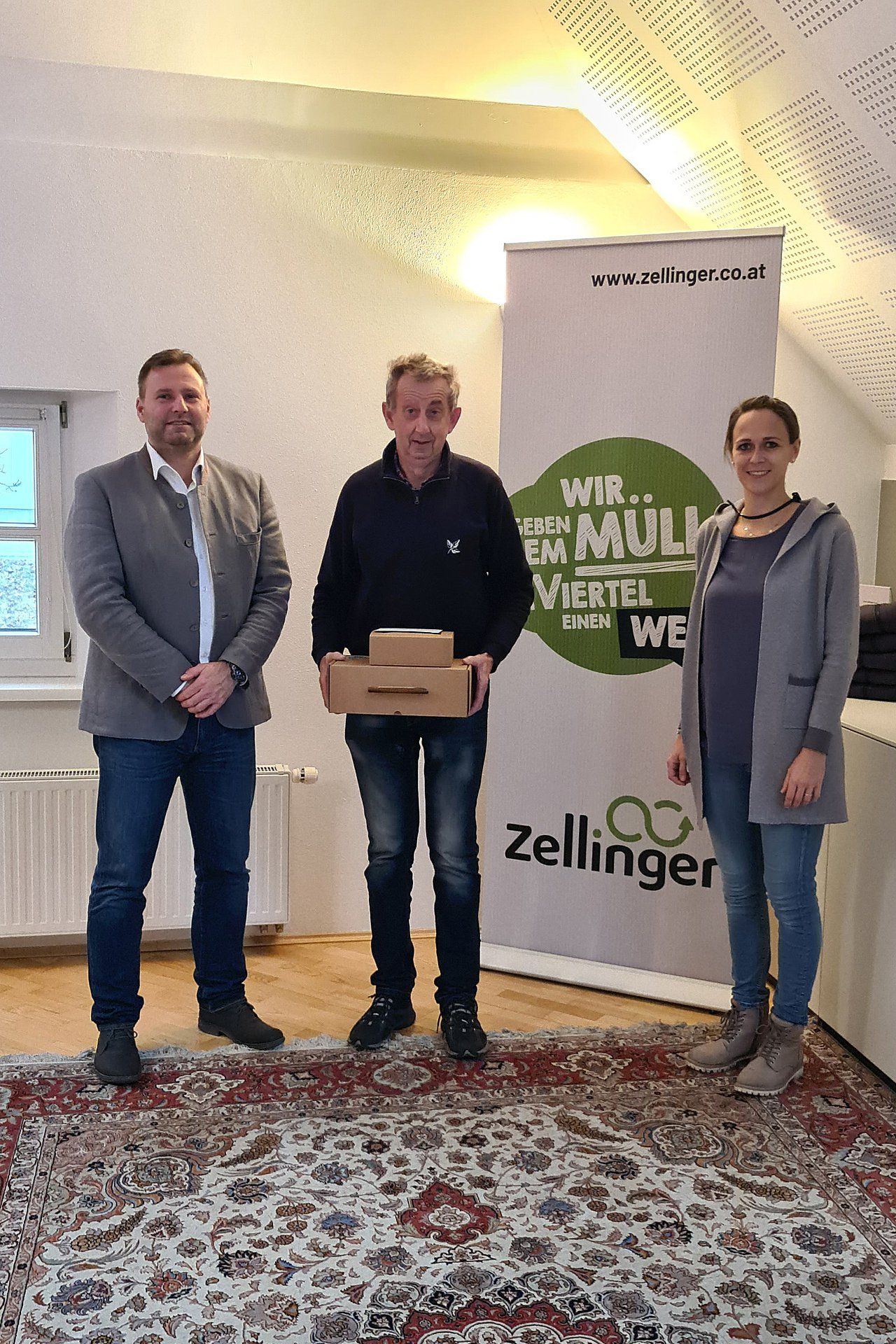 Three people standing; man holding boxes in front of a banner with logo; inside room with rug.