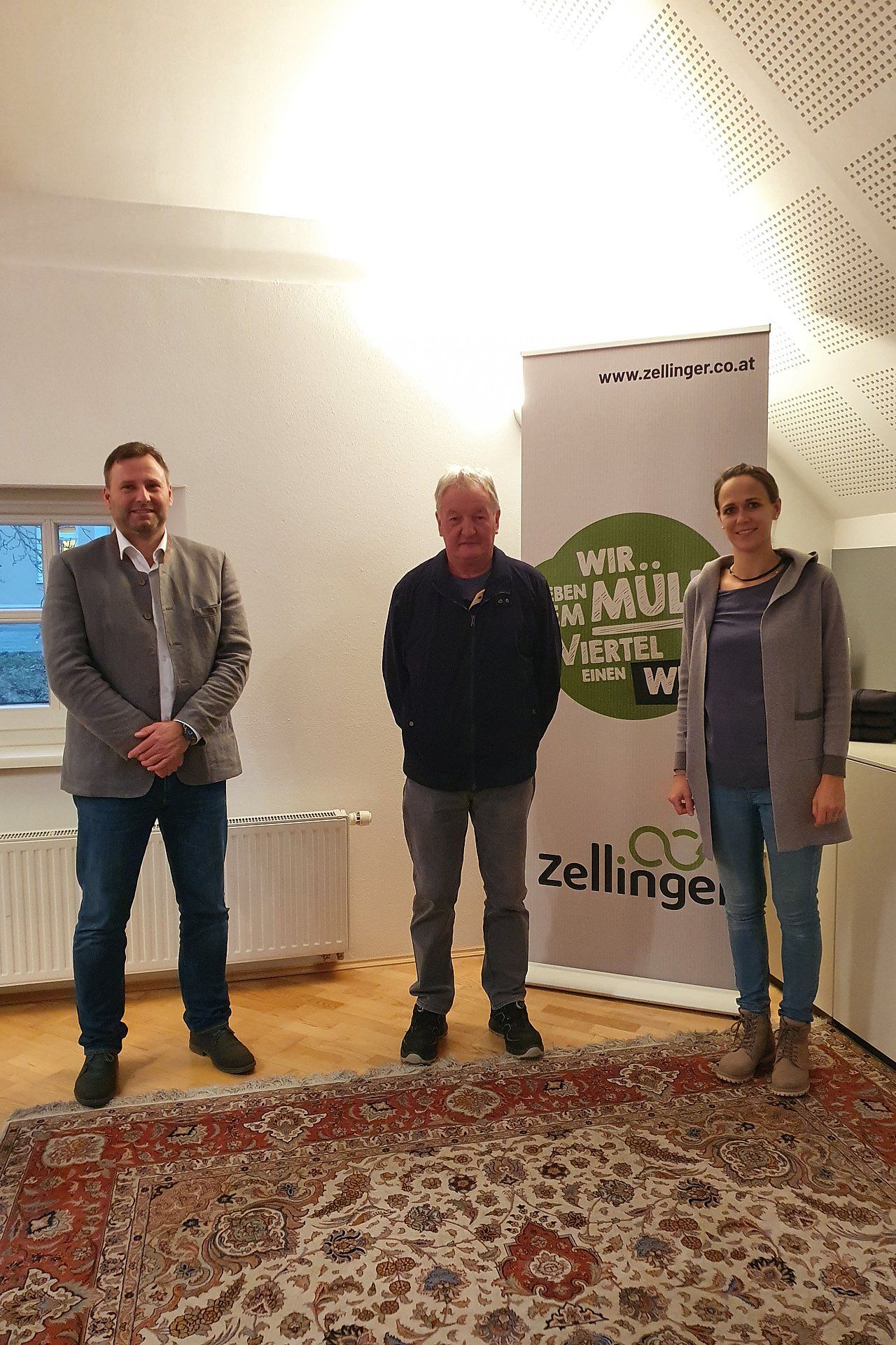 Three people standing, posing indoors in front of a banner with a logo.