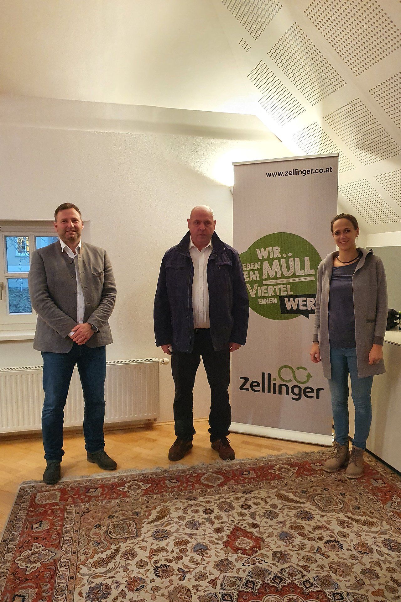 Three people stand indoors near a banner. A man in a jacket and jeans stands between two others.