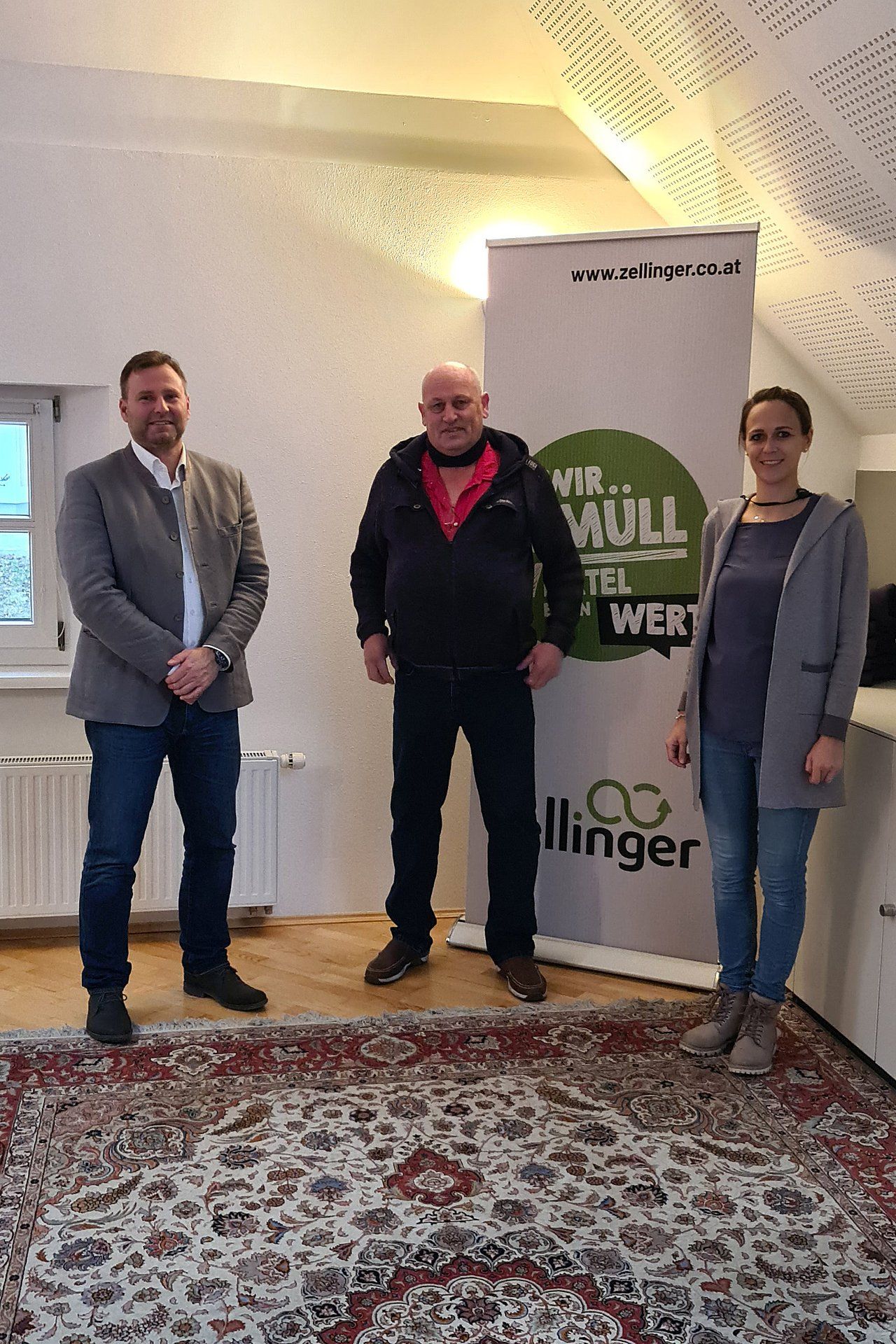 Three people standing, posing in front of a banner; a man in a blazer, a man in a jacket, and a woman in a hoodie.