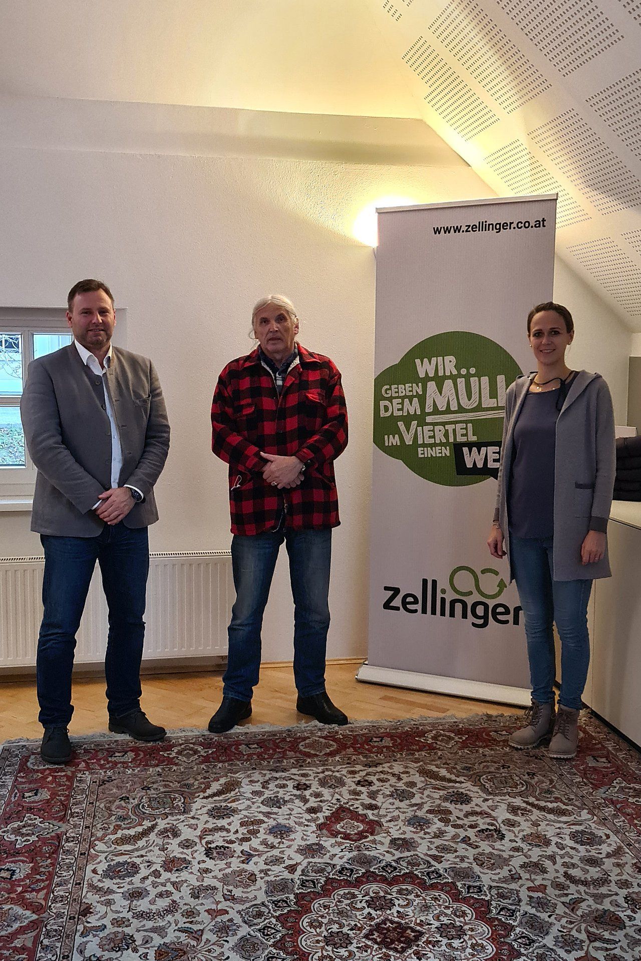 Three people stand indoors near a banner. A man in a red plaid shirt stands between two others.