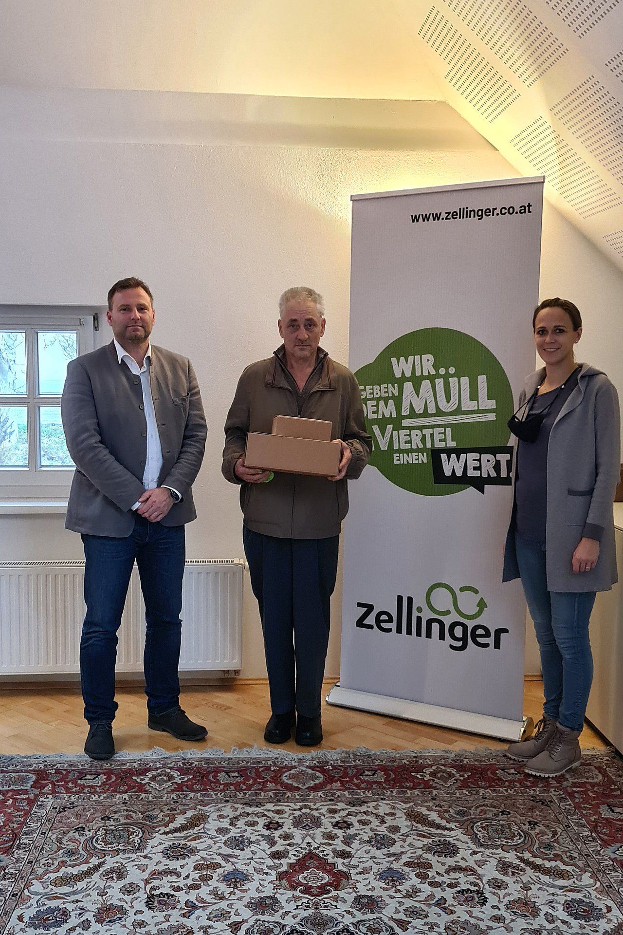 Three people stand indoors holding cardboard boxes in front of a banner.