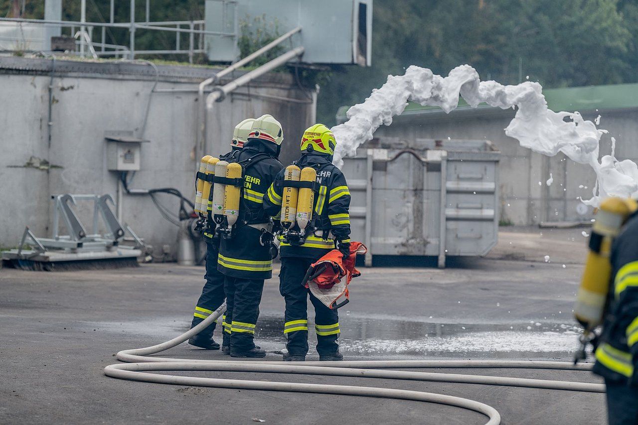 Firefighters in protective gear using foam to extinguish a fire near a building.
