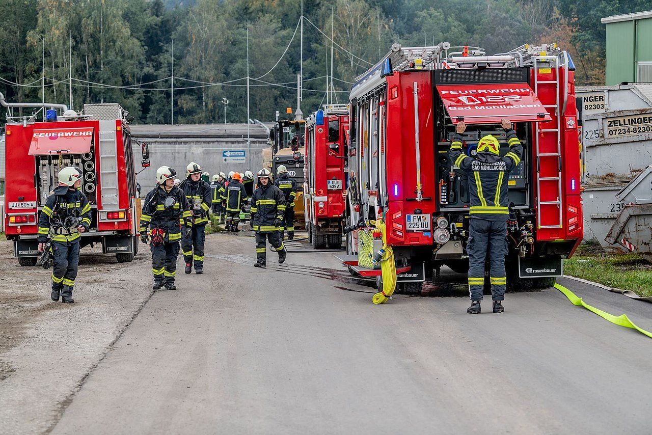 Firefighters and fire trucks at a scene. Several firefighters walk toward the trucks. Red fire trucks and equipment are present.