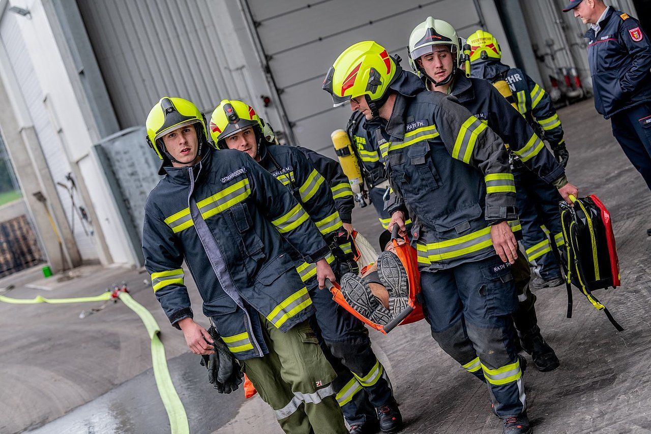 Firefighters in yellow helmets and jackets carry a stretcher, likely during a rescue operation at a fire station.