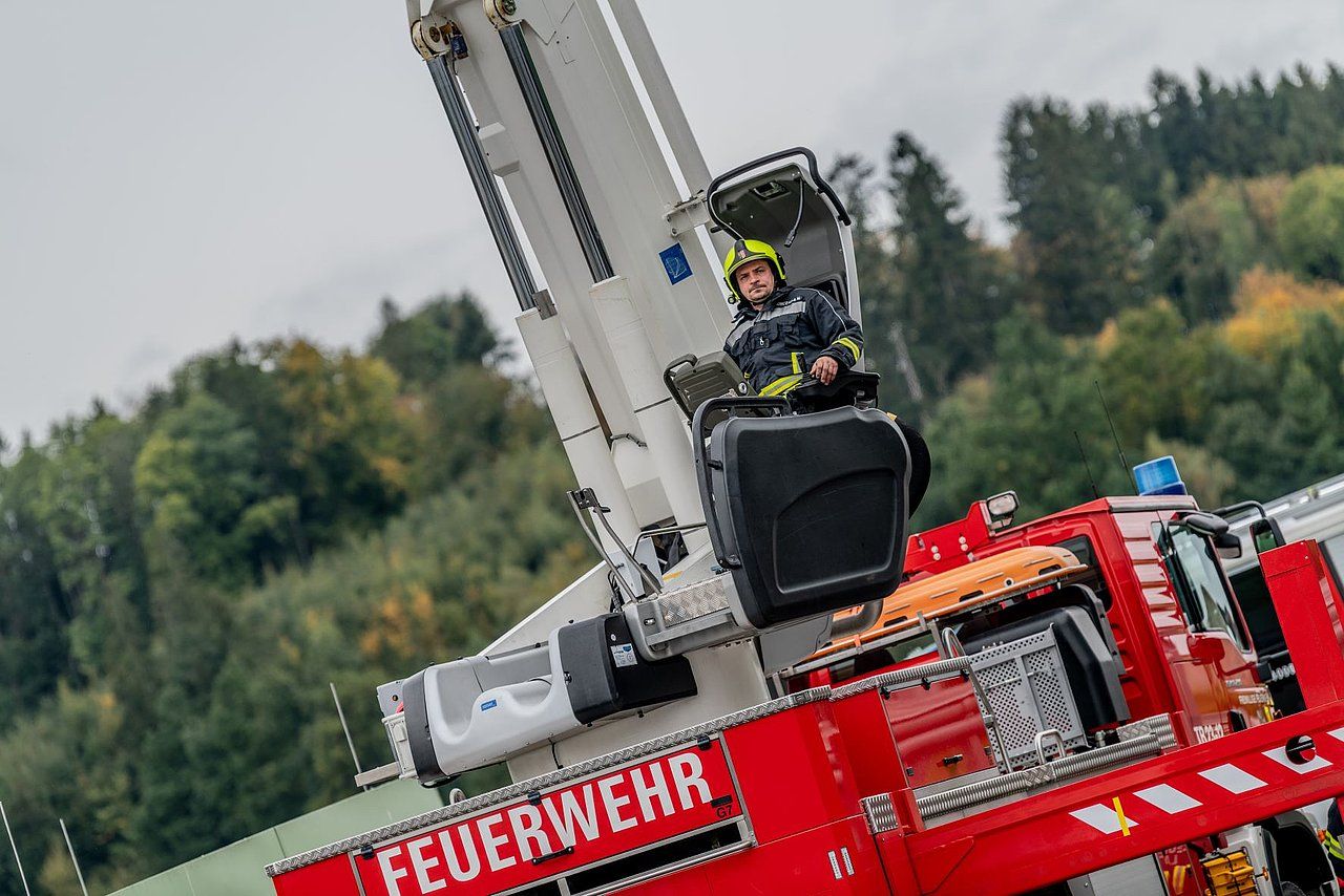 Firefighter in a red truck's extended aerial platform, raised against a forest backdrop.