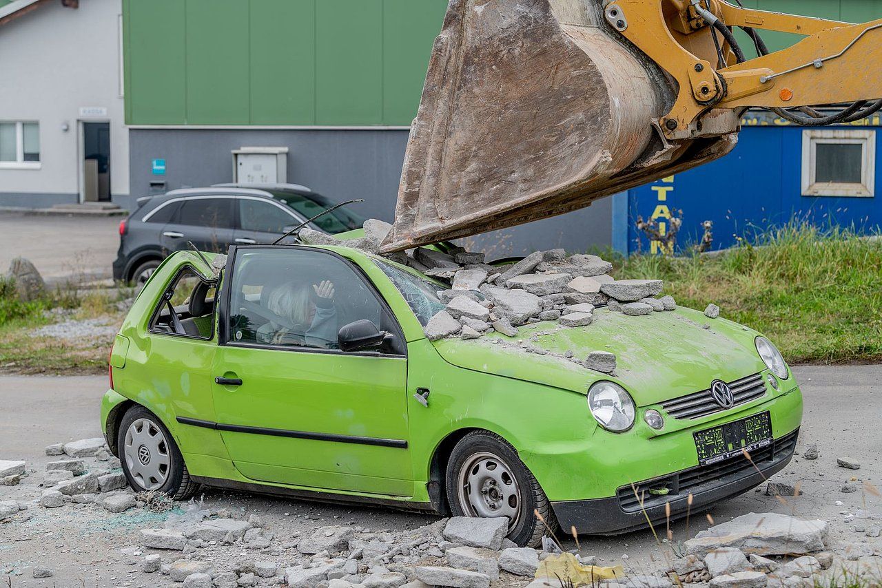 Green car being crushed by an excavator bucket filled with concrete debris. Outdoors.