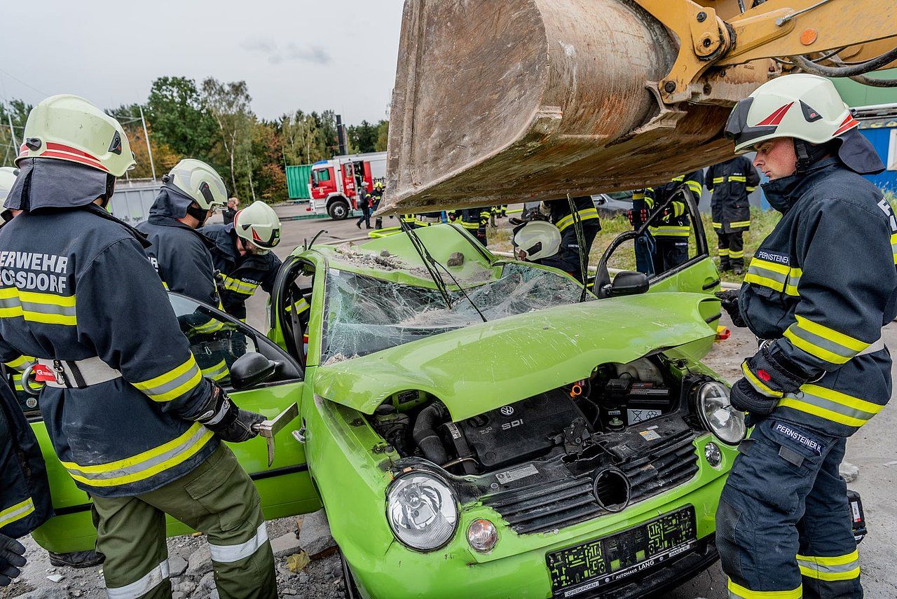 Firefighters using heavy machinery to extract a person from a crushed green car at a rescue training.