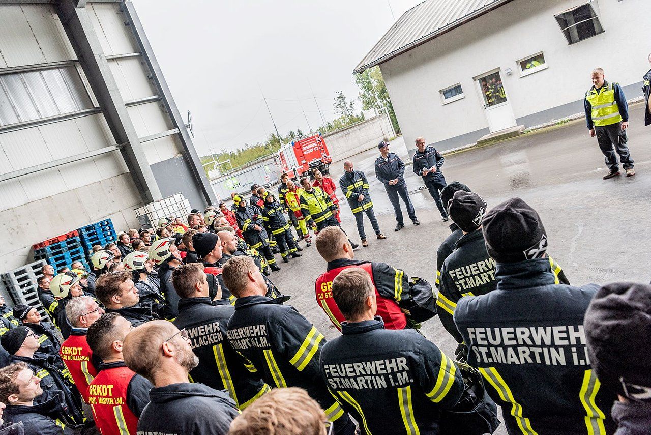 Firefighters in black and yellow uniforms gathered outdoors, some looking upward; a building and sky are in the background.