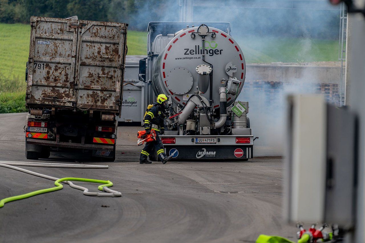 Firefighters at the rear of a tanker truck, smoke visible. Dump truck in the background. Outdoors.