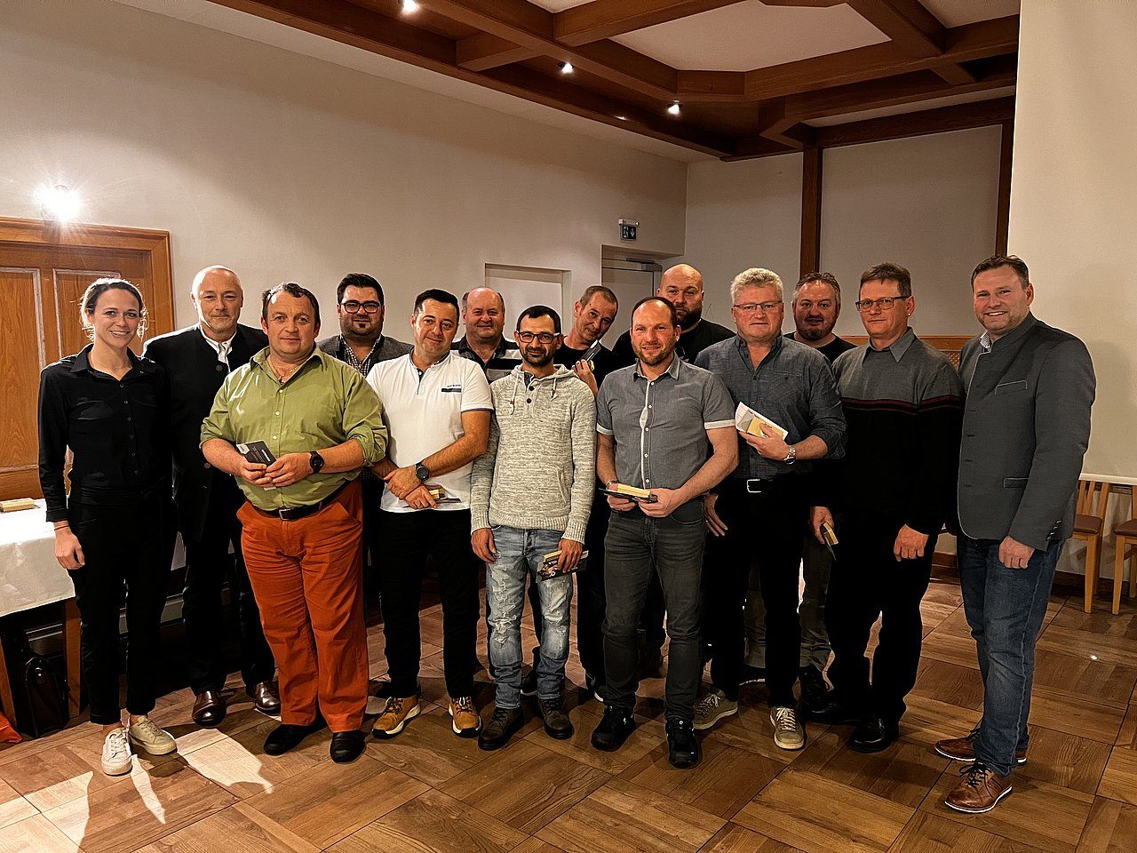 Group of people smiling in a room, possibly an event. Men and one woman in varied attire. Wooden walls and ceiling.