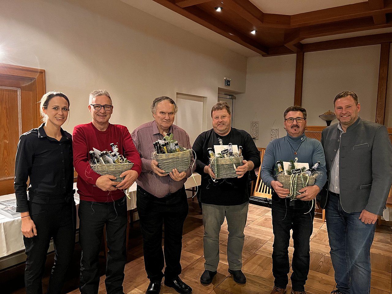 Six people in a room, holding gift baskets. Men wear casual clothes, one with a jacket. They appear to be at an event.