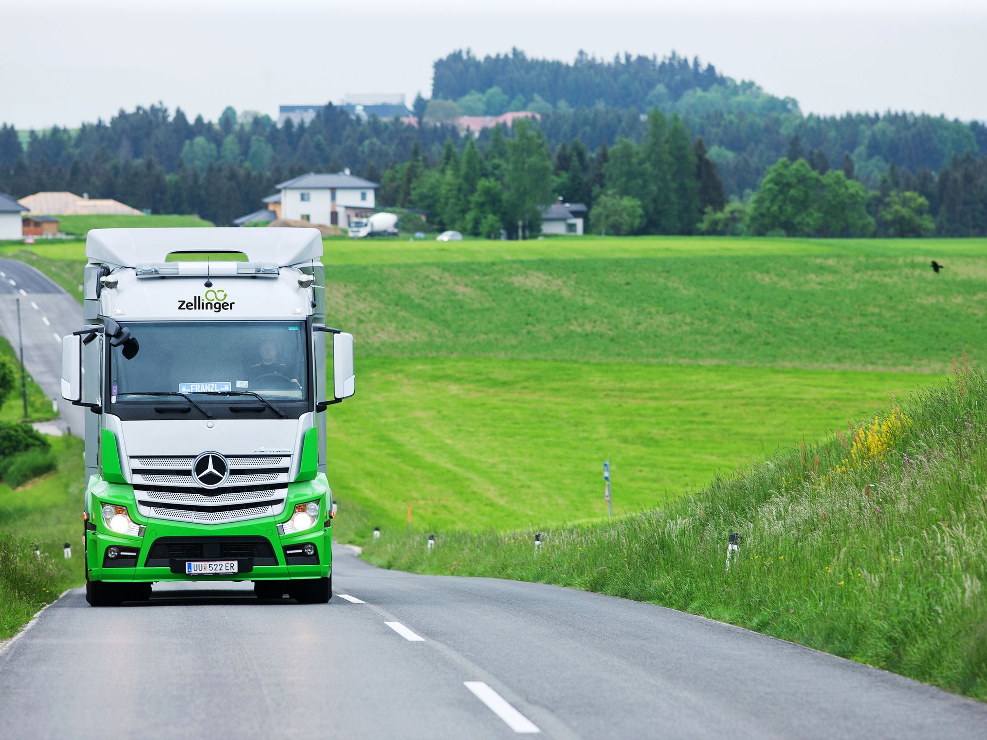 Green and silver semi-truck driving on a road, surrounded by green fields and trees.
