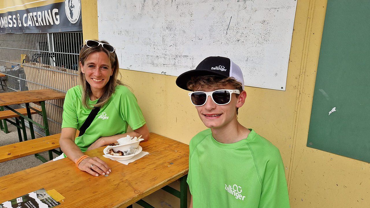 Woman and teen in green shirts, seated at a picnic table outdoors. Smiling, they wear sunglasses and a baseball cap.