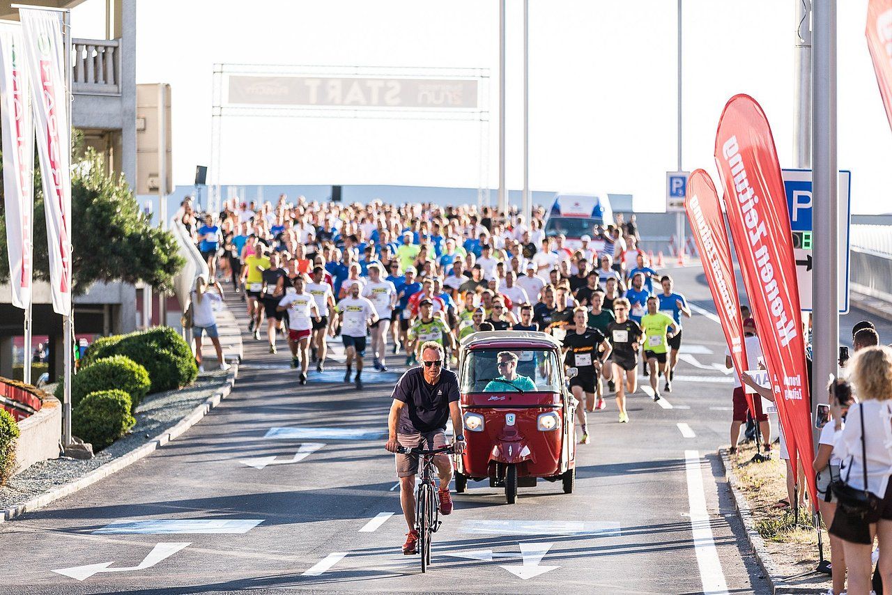 Runners in a road race with a cyclist in front and a tuk-tuk behind. Bright day.