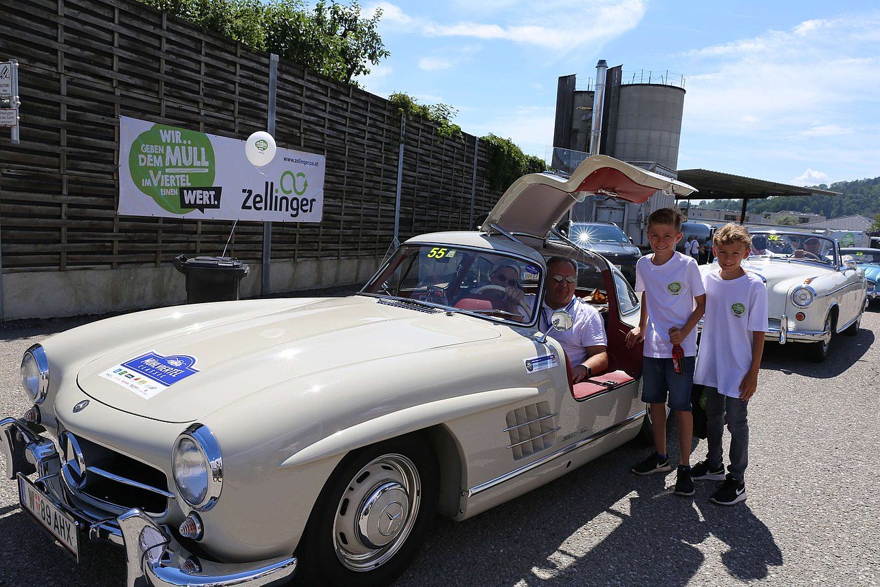 Man in vintage car with gull-wing doors, poses with two boys. Outside, near a sign and other classic cars.
