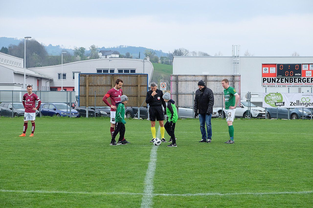 Soccer players and referee on field before game, green and maroon team, cars and buildings in background.