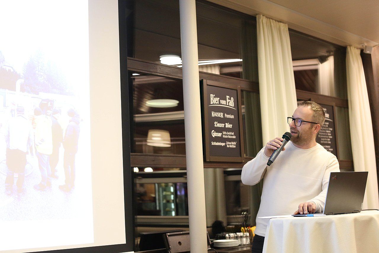 Man in a white sweater speaking at a presentation. A projector screen displays a photo, and a laptop sits on a table.