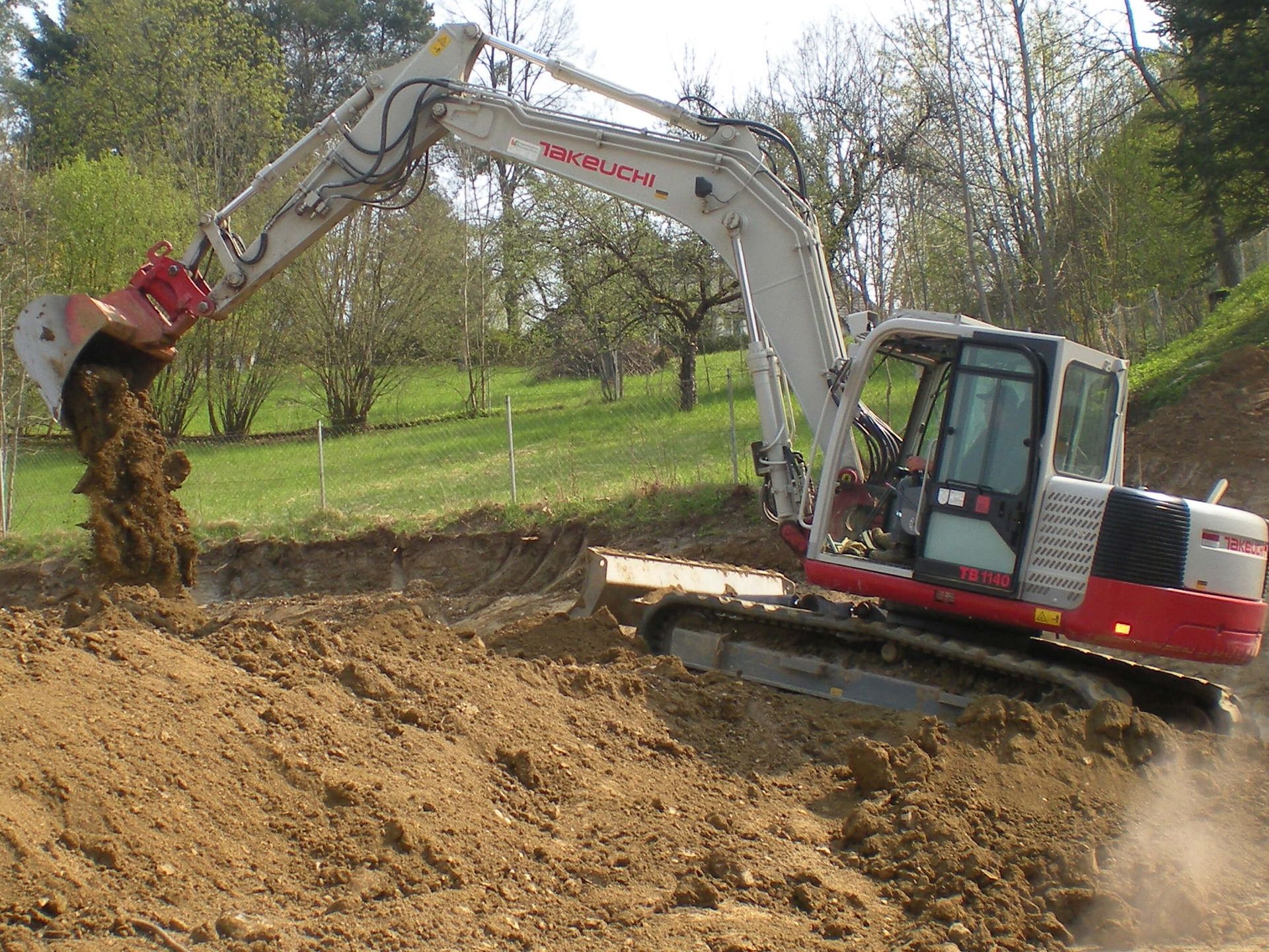 Excavator digging earth in a construction site; brown soil and green background with trees.