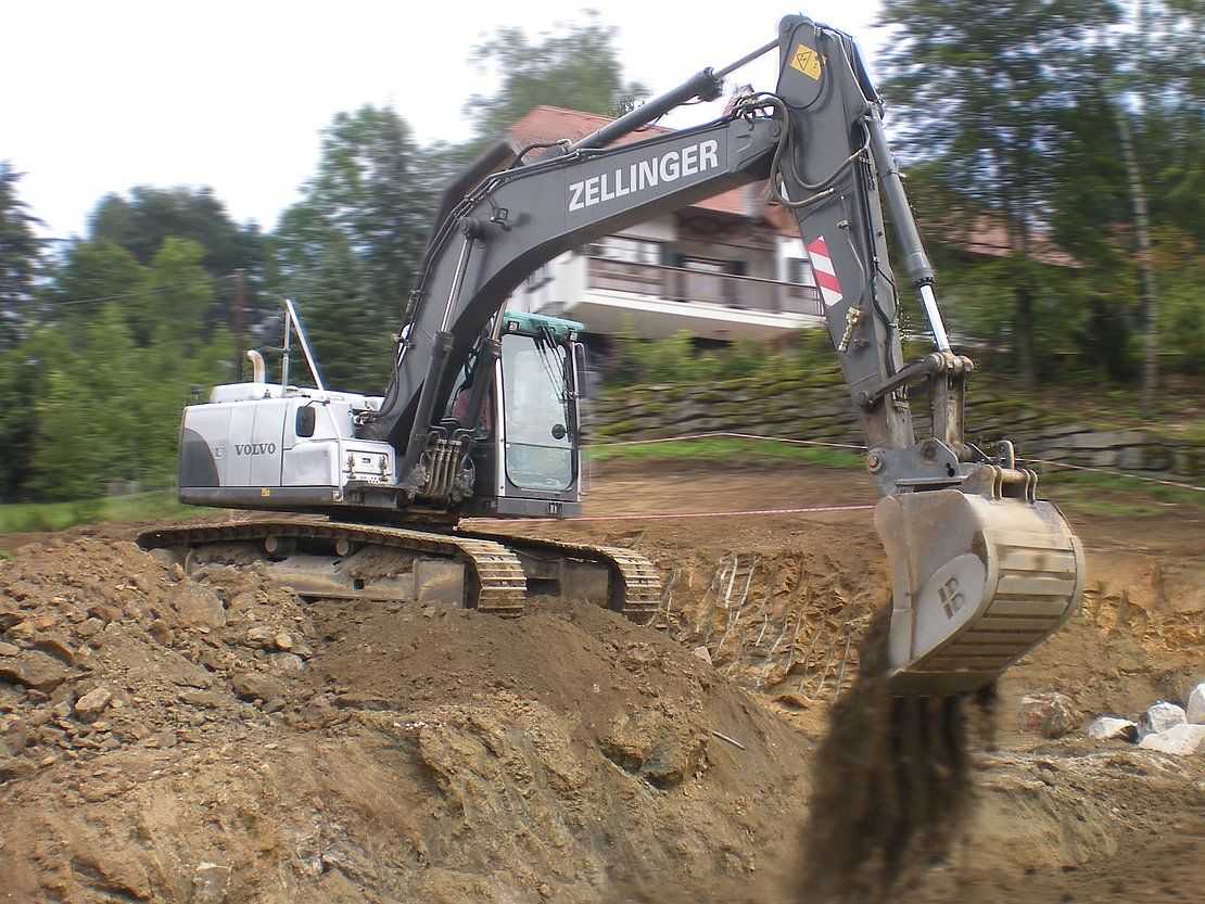 Excavator digging into a hillside, scooping dirt. Gray machine with black details, outdoors in a wooded area.