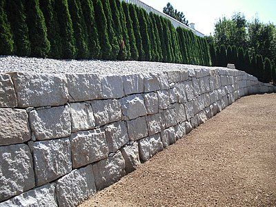 Stone retaining wall with gravel path, topped with white rocks and green hedge.