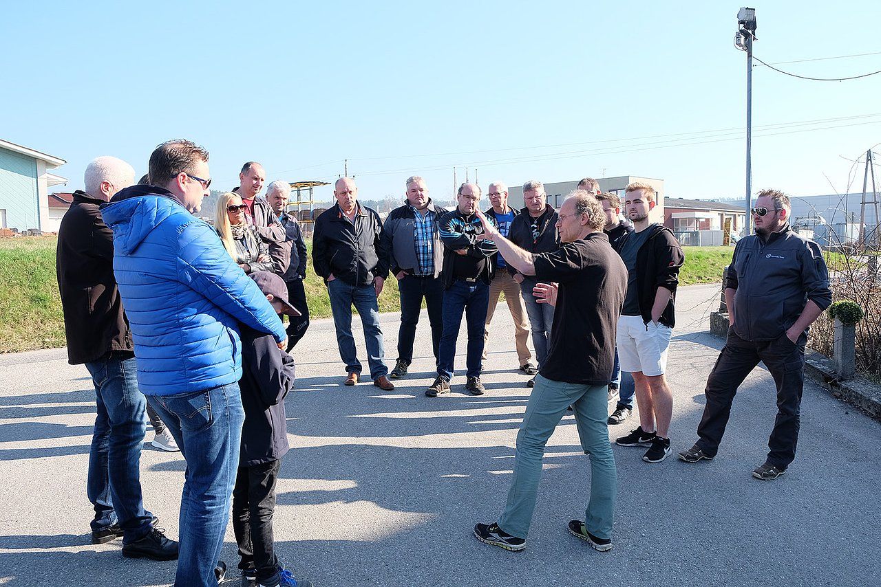 Group of people outdoors listening to a woman gesturing, sunny day, light blue sky.