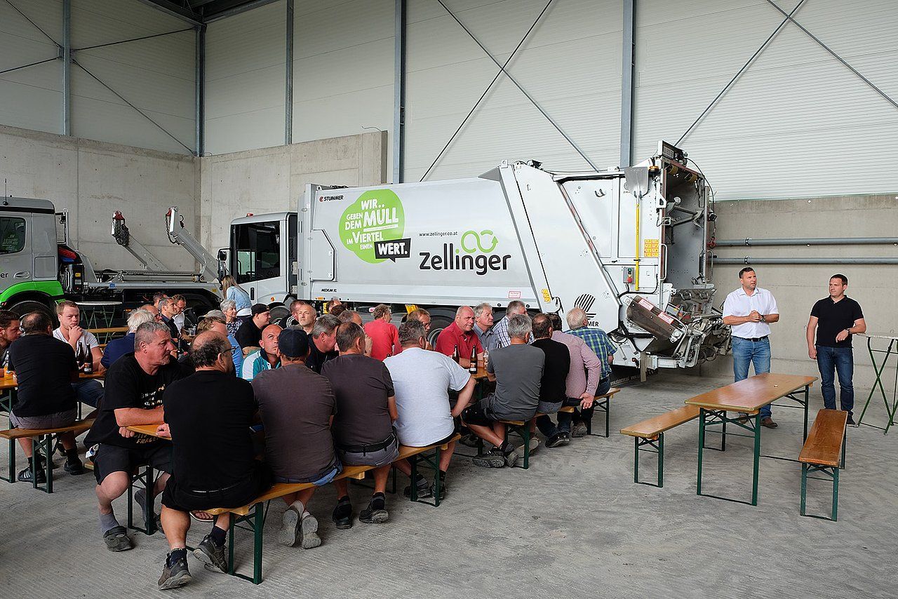 Group of people at tables, likely a gathering, with a garbage truck in a shed.