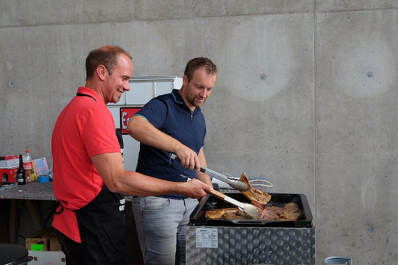 Two men grilling steaks outdoors, one in red shirt smiling.