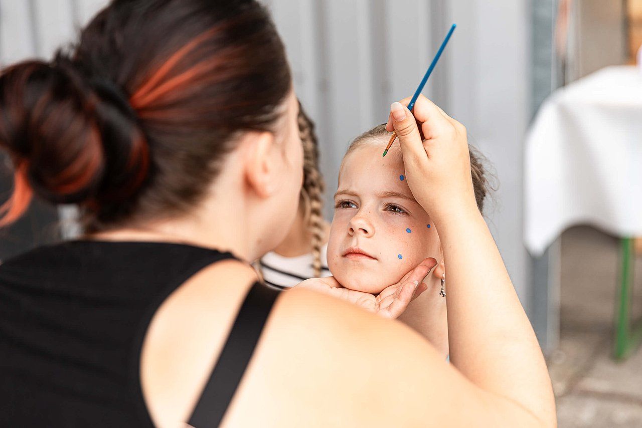 Woman painting glitter on a child's face outdoors; a sunny day, focusing on the child's face and artist's hand.
