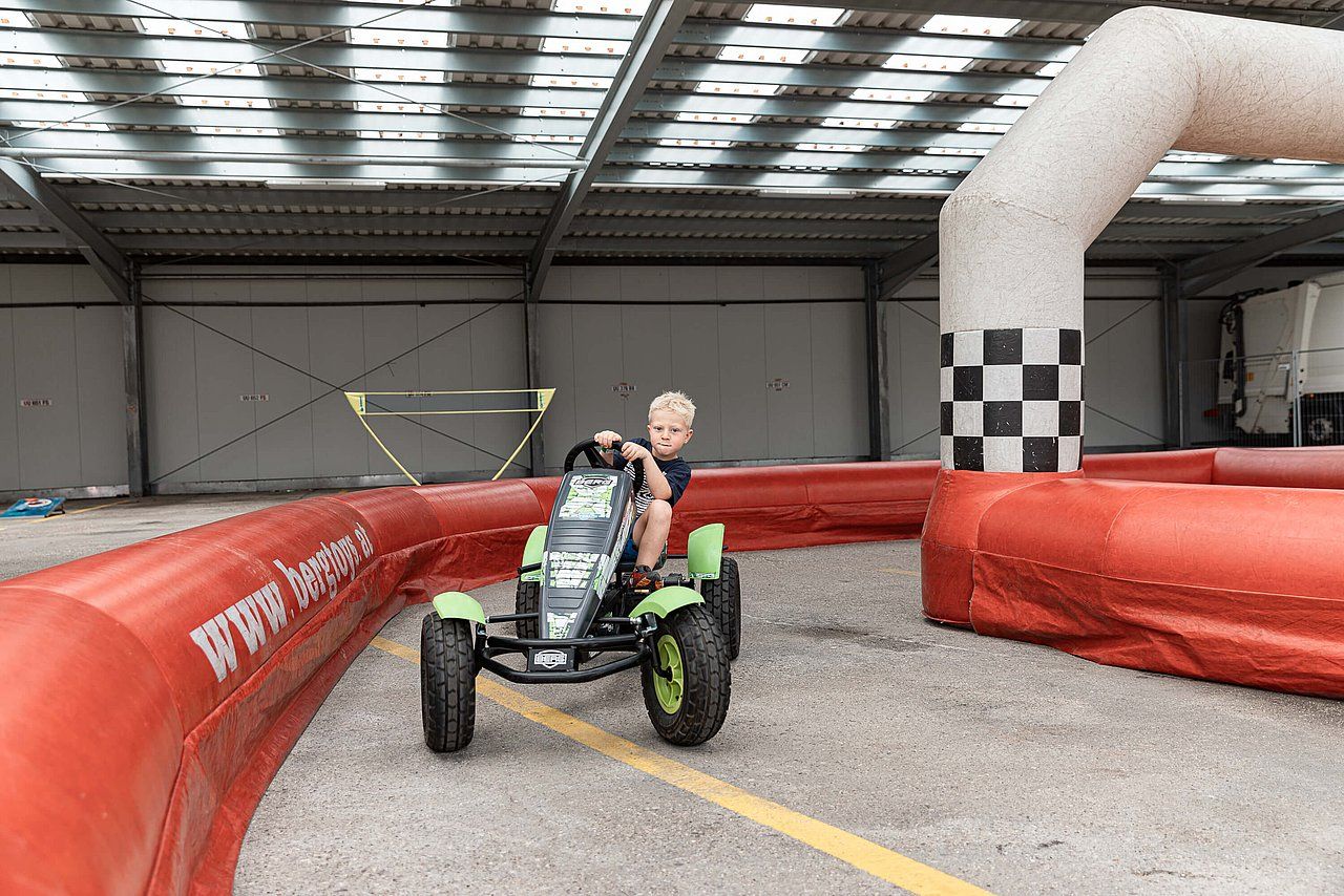 Boy driving a green pedal go-kart on a small track with red barriers and a checkered archway.