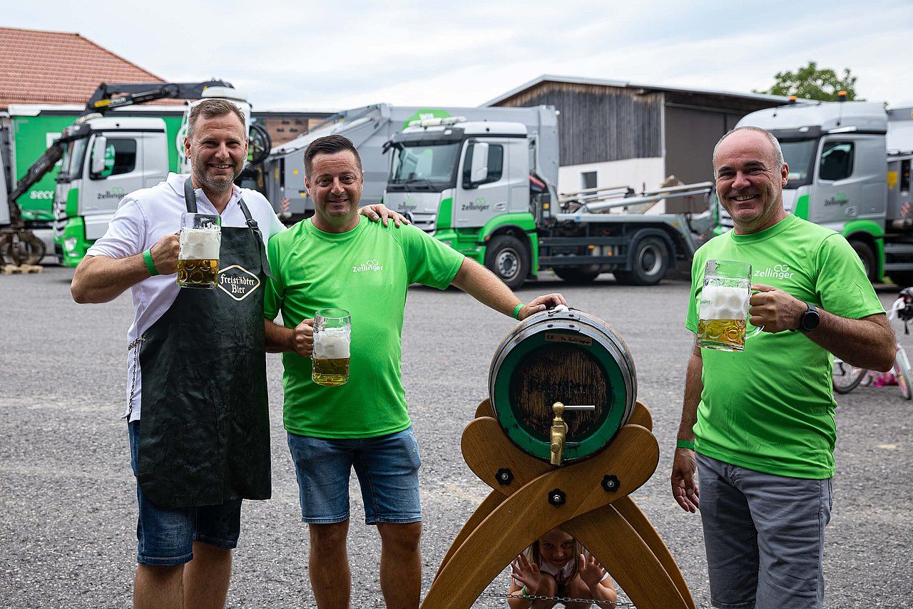 Three men with beer mugs beside a keg, in front of green trucks.