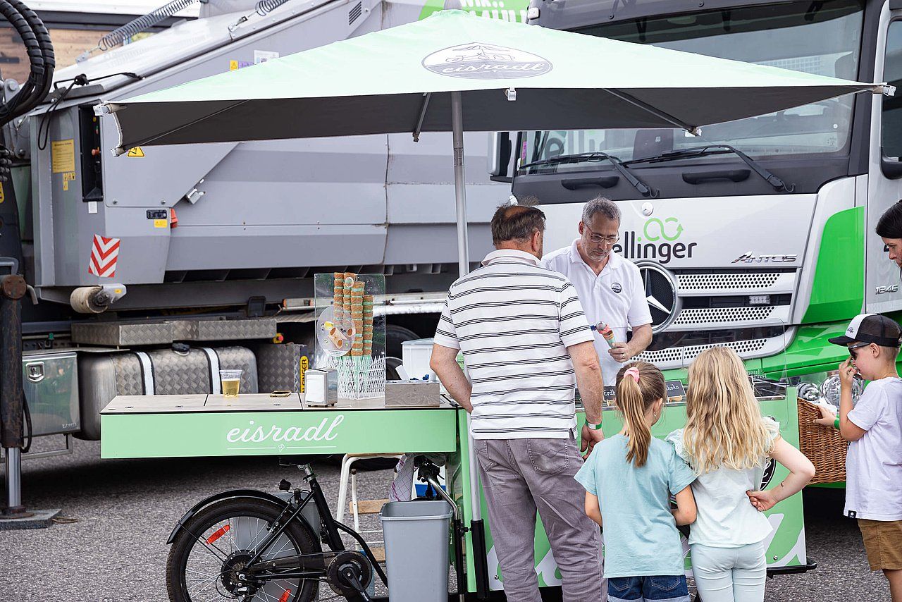 A food stand with customers, next to a green truck. People are gathered under a green umbrella.