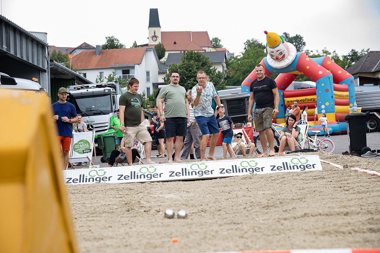 Men playing petanque in a sandy area, with a clown-themed bouncy castle and buildings in the background.