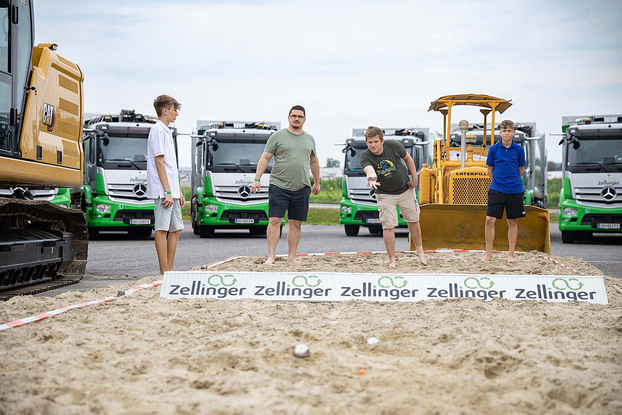 Four men and a boy playing boules in a sandy area, with construction vehicles in the background.