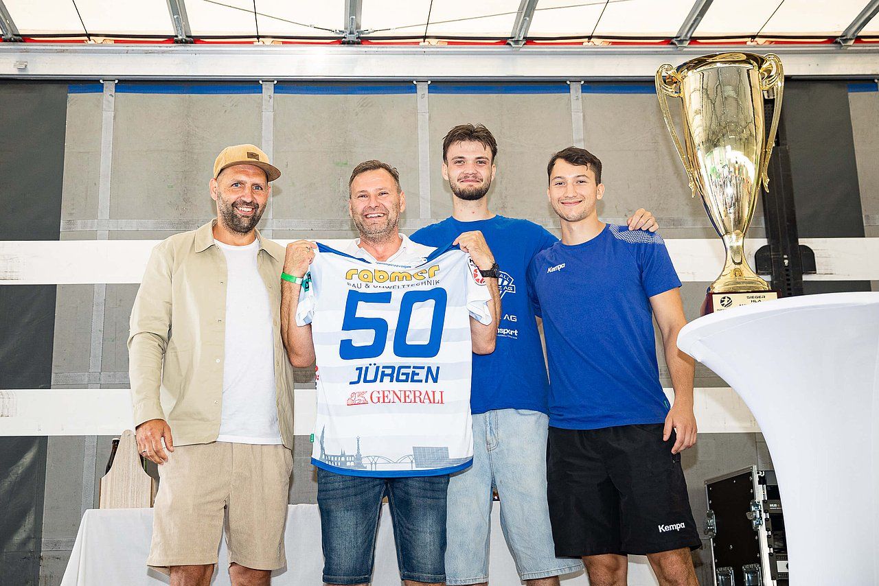 Four men pose with a trophy, one holding a jersey. They stand outdoors; the jersey is blue, white, and black.
