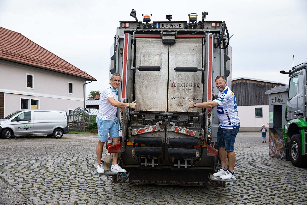 Two men standing on the back of a garbage truck, pointing. Cloudy outdoor setting.