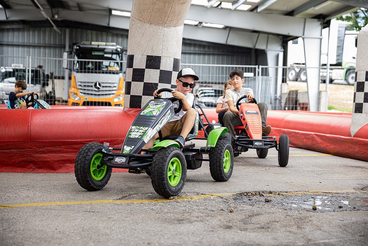 Two boys on pedal go-karts at an outdoor track, green and orange vehicles, checkered finish line.