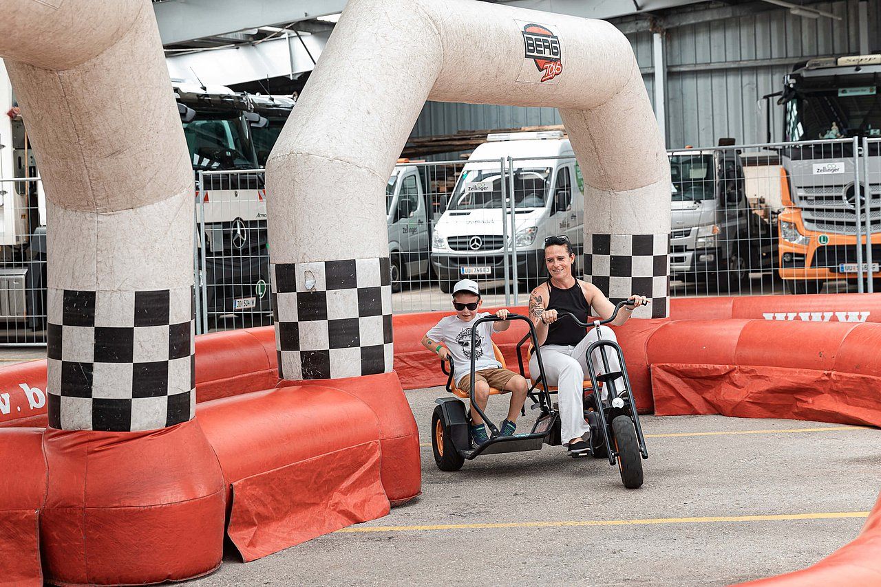 Two people on a trike smile and wave under a checkered archway at a go-kart event.