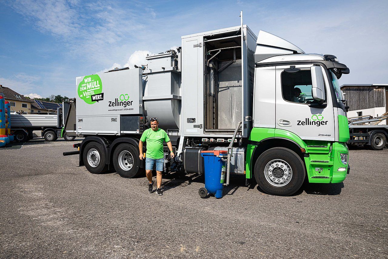 Man in green shirt by a recycling truck. Truck is silver and green. Outdoor setting.
