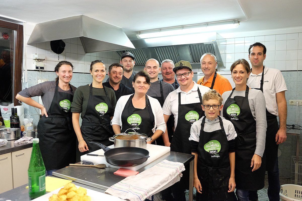 Group of people in a kitchen wearing aprons, smiling at the camera, next to cooking equipment.