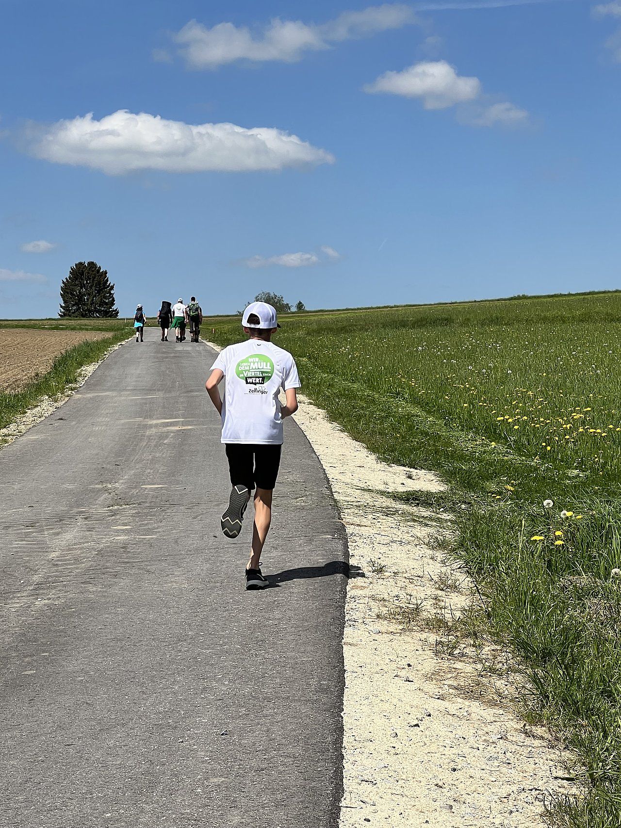 Runner on a path, others ahead. Bright blue sky, fields of green and yellow flowers, sunny day.