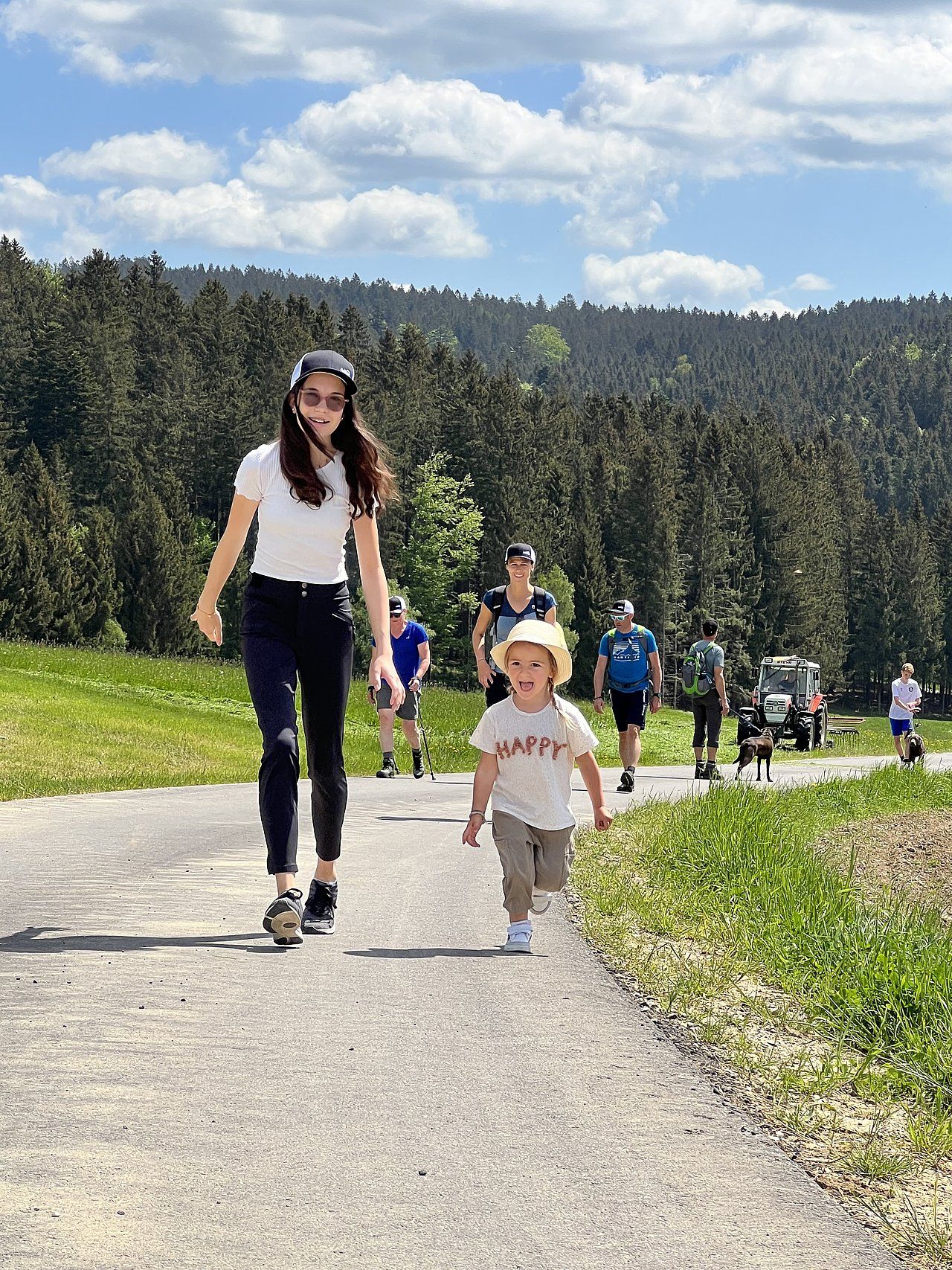 Woman and toddler walking on a path, followed by other people. Green field, blue sky, and forest in the background.