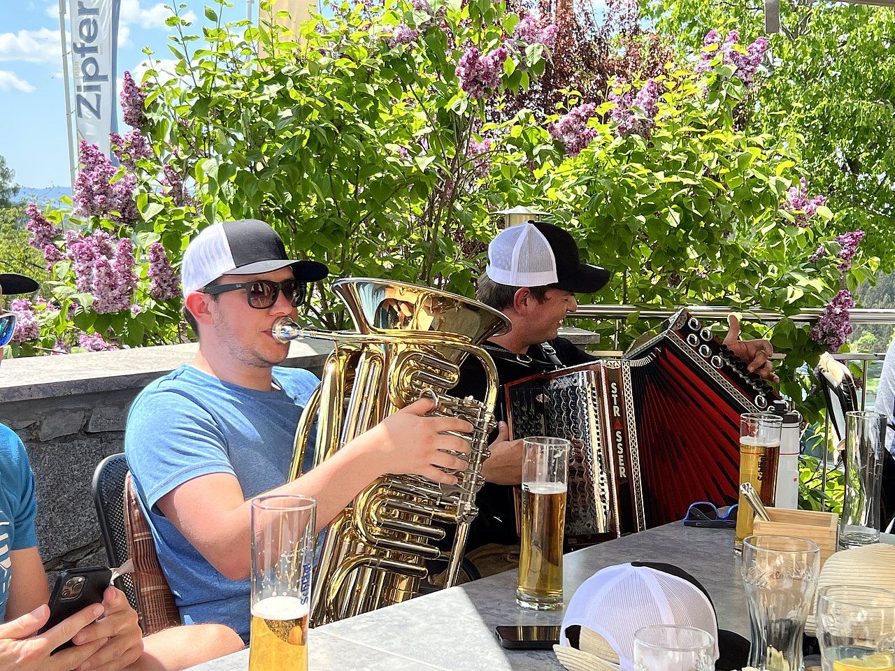 Two men playing music outdoors, one a tuba, the other an accordion. They are at a table with beer.