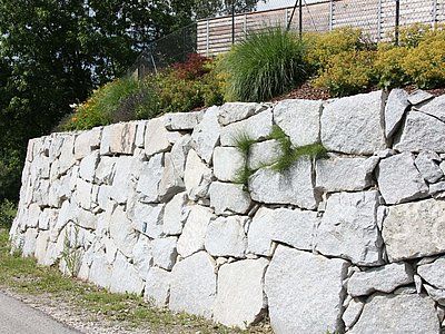 Stone retaining wall supporting a hillside garden with plants and grass.