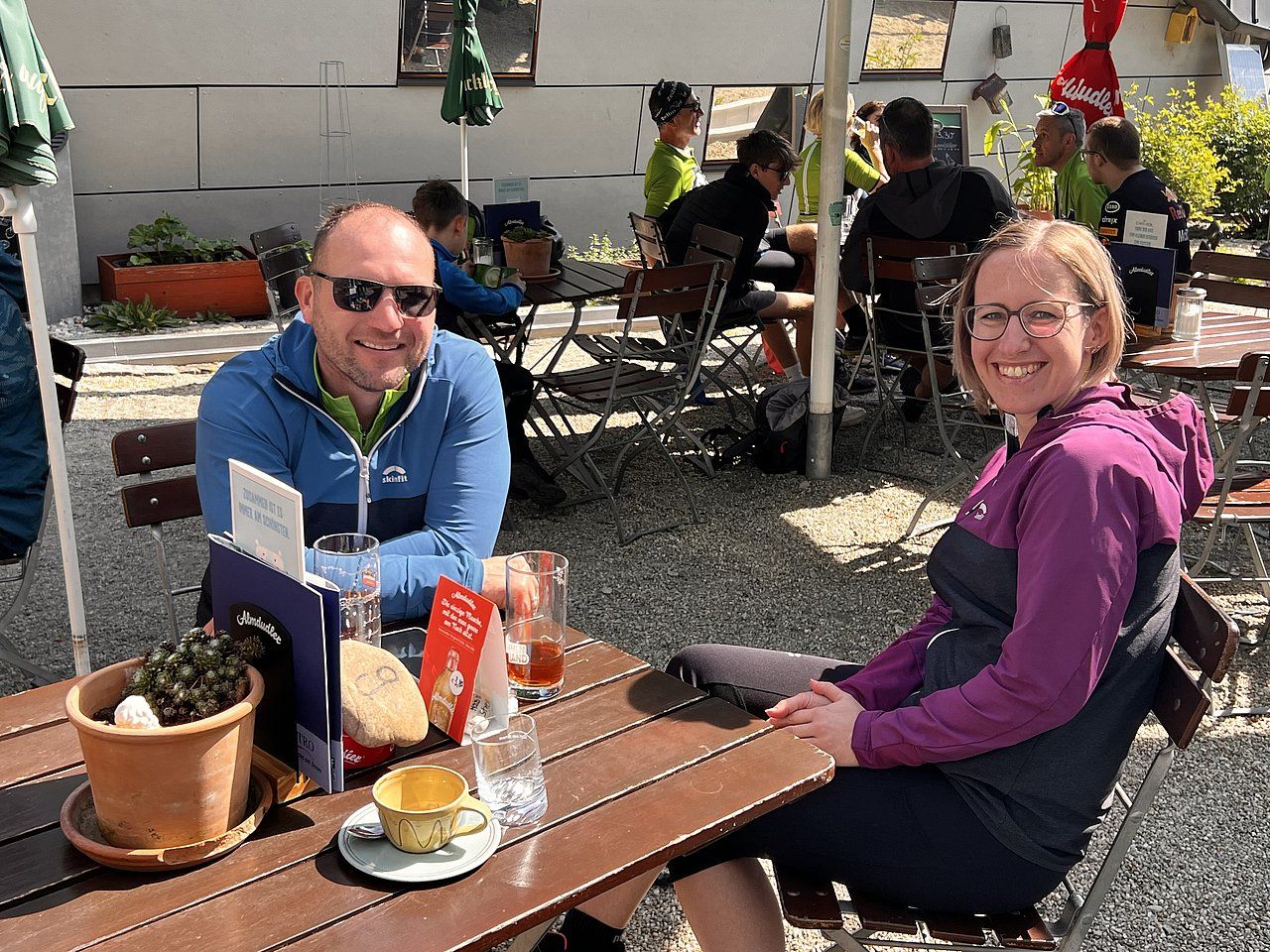 Two people seated at a table outdoors. Man smiles, wearing blue. Woman smiles, wearing purple. Other people nearby.