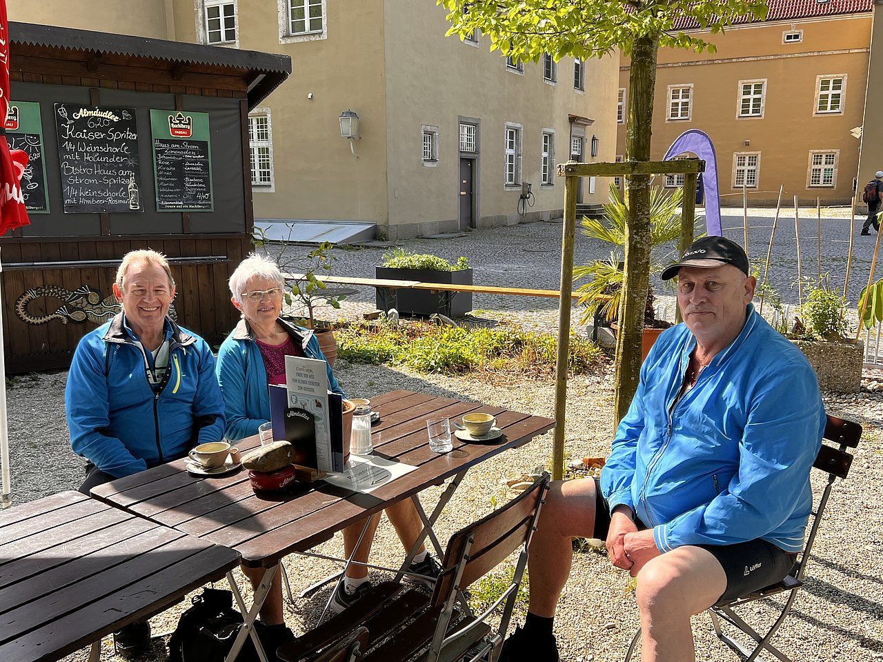 Three people in blue jackets sit at an outdoor cafe table. Buildings and plants in background.