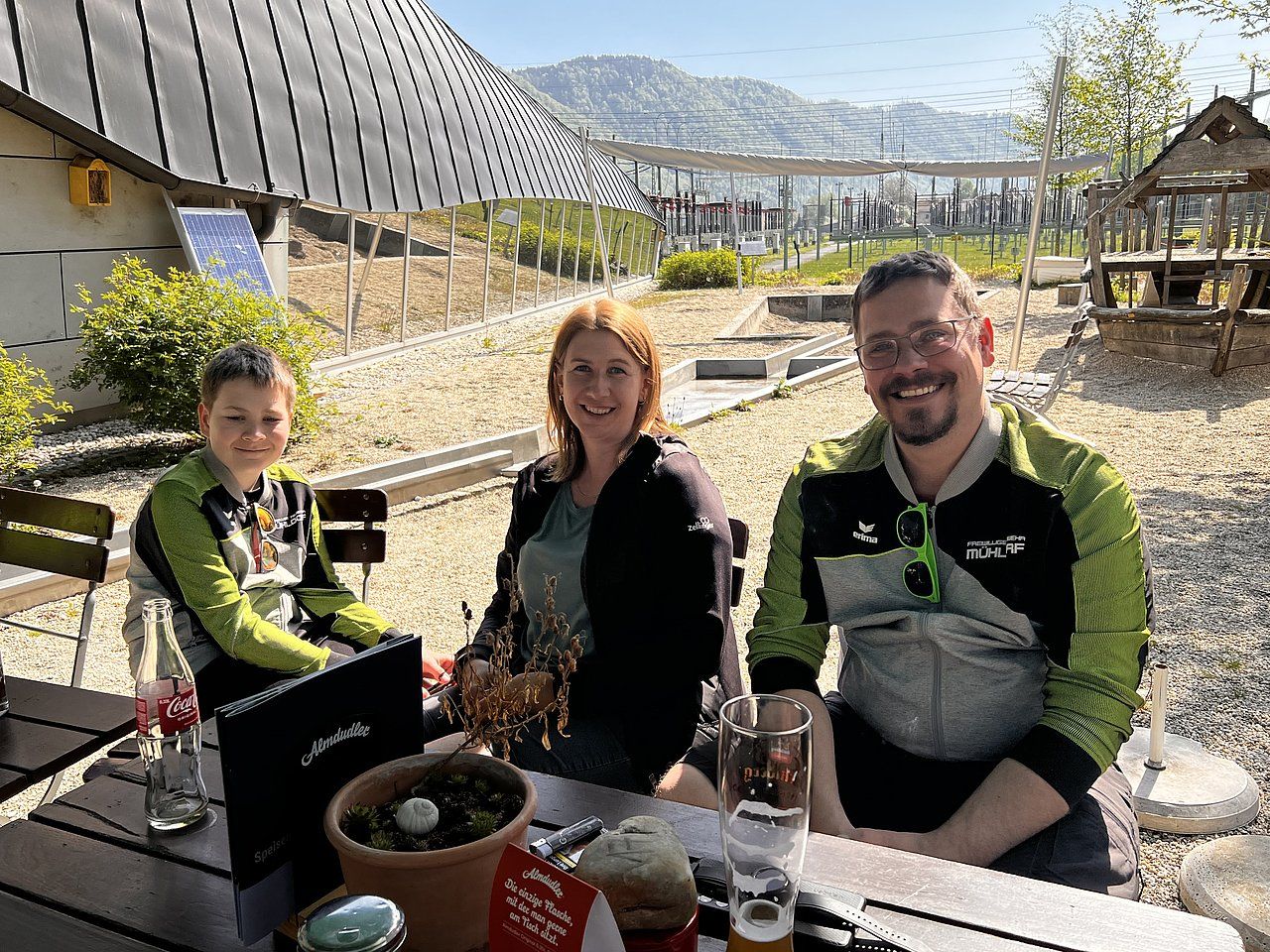 Three people smiling at an outdoor cafe table. A green and gray mountain view background.
