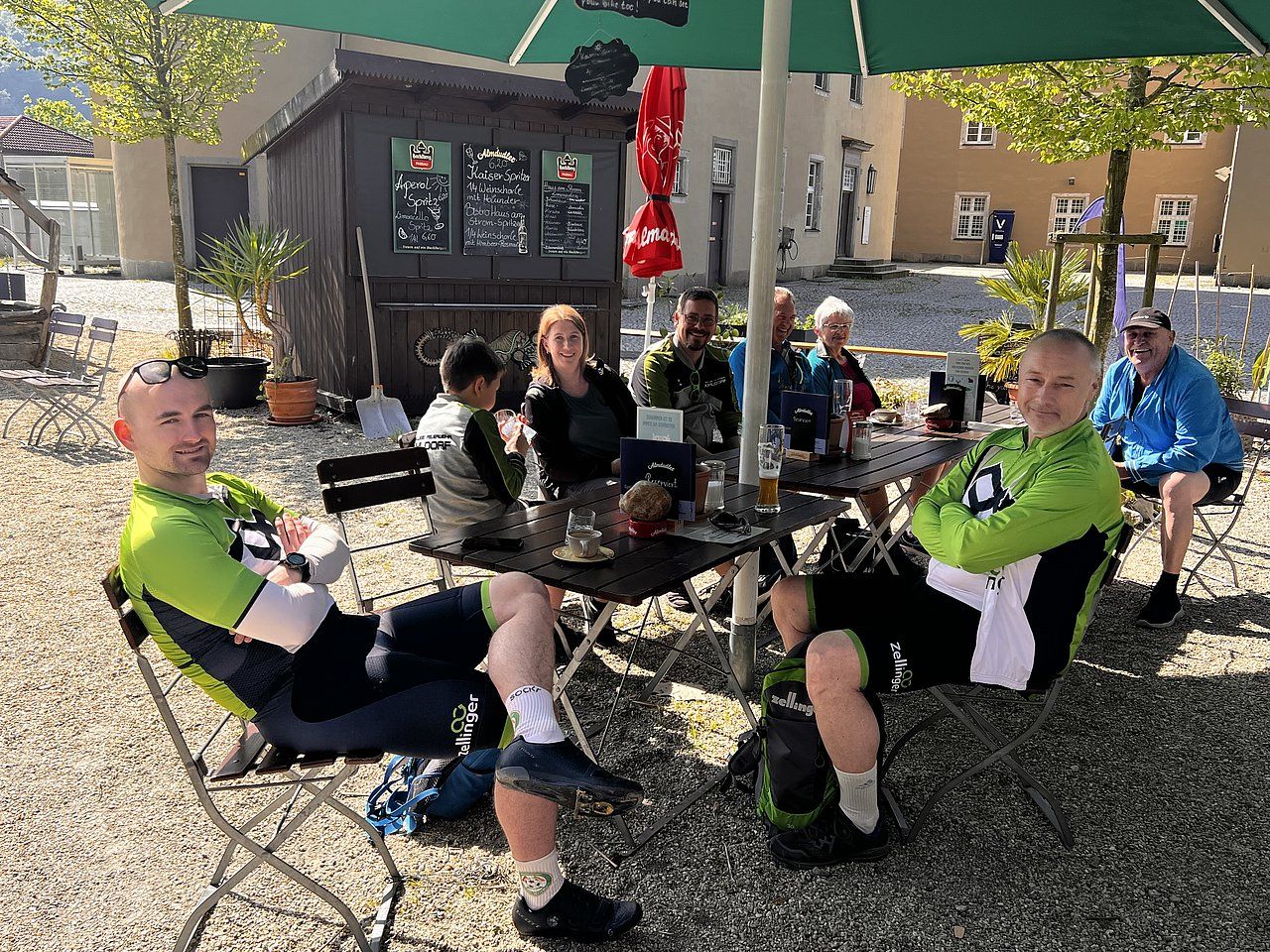 People relaxing at an outdoor cafe under a green umbrella; two cyclists sit in foreground.