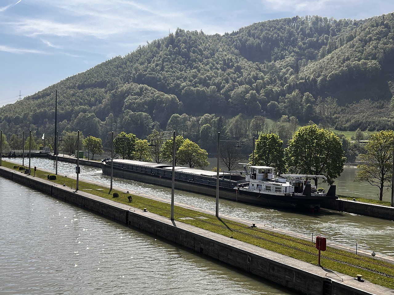 A cargo barge travels through a canal bordered by green grass and trees, with a forested hillside in the background.