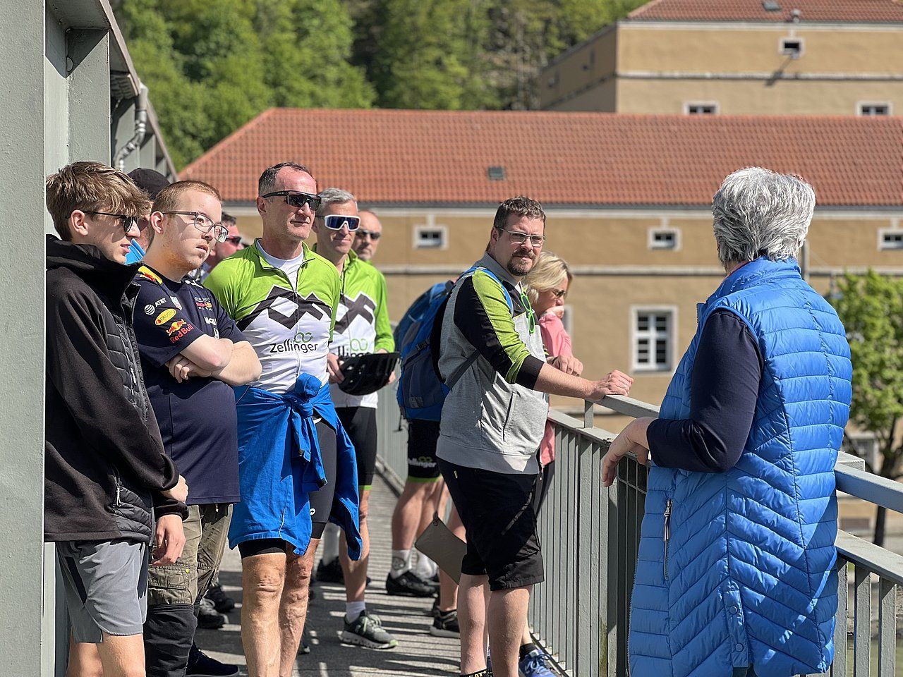 Group of people on a bridge, listening to a woman. Historic building visible.